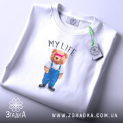 Футболка моє життя з ведмедиком, біла, складена, фон світло-фіолетовий, принт ведмедика в синіх штанях і білу футболку з написом 'MY LIFE', на етикетці зелена стрічка.