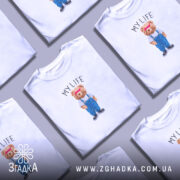 Футболка моє життя з ведмедиком, біла, на моделі, фон світло-фіолетовий, принт ведмедика в синіх штанях і білу футболку з написом 'MY LIFE'.