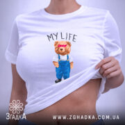 Футболка моє життя з ведмедиком, біла, складена, фон світло-фіолетовий, принт ведмедика в синіх штанях і білу футболку з написом 'MY LIFE', на етикетці зелена стрічка.