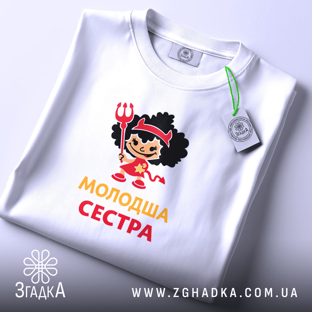 Футболка молодша сестра, біла, на столі, з етикеткою, фон світло-сірий, принт з дівчинкою в червоному вбранні з чорними кучерями, тримає тризубець, текст 'молодша сестра' жовтого кольору.