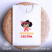 Футболка молодша сестра, біла, на плетеній основі, фон білий, принт з дівчинкою в червоному вбранні з чорними кучерями, тримає тризубець, текст 'молодша сестра' жовтого кольору.