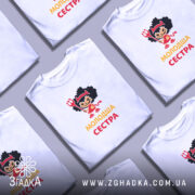 Футболка молодша сестра, біла, на моделі, з легким макіяжем, фон світло-фіолетовий, принт з дівчинкою в червоному вбранні з чорними кучерями, тримає тризубець, текст 'молодша сестра' жовтого кольору.