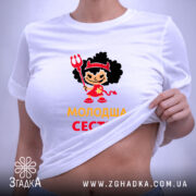 Футболка молодша сестра, біла, на столі, з етикеткою, фон світло-сірий, принт з дівчинкою в червоному вбранні з чорними кучерями, тримає тризубець, текст 'молодша сестра' жовтого кольору.