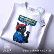 Футболка ми можемо це зробити, біла, на столі, фон світло-сірий, принт з чорним котом у червоній бандані, що піднімає кулак, текст на футболці синій з жовтим, з етикеткою.