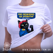 Футболка ми можемо це зробити, біла, на столі, фон світло-сірий, принт з чорним котом у червоній бандані, що піднімає кулак, текст на футболці синій з жовтим, з етикеткою.