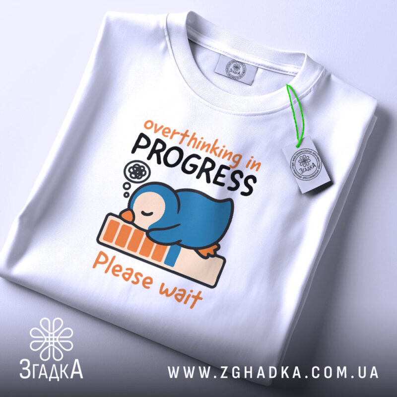 Футболка мислення в процесі: біла футболка, складена на поверхні, з принтом пінгвіна, що спить на барі прогресу, фон світло-сірий, деталі принта чіткі, текст 'overthinking in PROGRESS Please wait'.