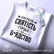 Футболка награна святість, біла, складена на поверхні, фон світло-сірий, принт чорного кольору з текстом на тему святості, з етикеткою зеленого кольору.