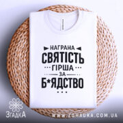 Футболка награна святість, біла, складена на кошику з ротанга, фон світло-сірий, принт чорного кольору з текстом на тему святості.