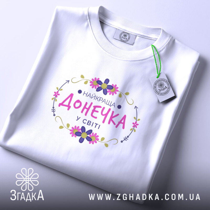 Футболка найкраща донечка у світі, біла, на столі, з етикеткою, фон світло-сірий, принт з написом 'НАЙКРАЩА ДОНЕЧКА У СВІТІ' в кольорах рожевий, фіолетовий та зелений, оформлений квітами.