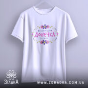 Футболка найкраща донечка у світі, біла, підвішена на вішалці, фон світло-сірий, принт з написом 'НАЙКРАЩА ДОНЕЧКА У СВІТІ' в кольорах рожевий, фіолетовий та зелений, оформлений квітами.