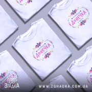 Футболка найкраща донечка у світі, біла, на моделі, з легким макіяжем, фон світло-фіолетовий, принт з написом 'НАЙКРАЩА ДОНЕЧКА У СВІТІ' в кольорах рожевий, фіолетовий та зелений, оформлений квітами.