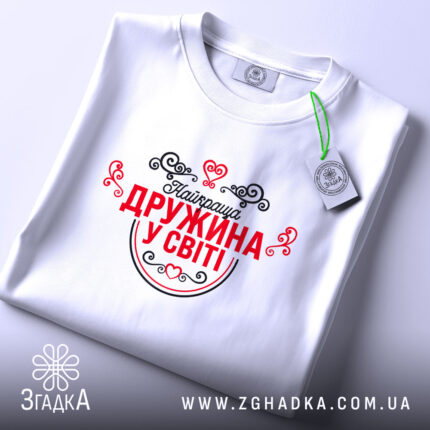 Футболка найкраща дружина. Біла футболка, складена на поверхні, зображена зверху. Принт червоного кольору з чорними елементами, напис "Найкраща дружина у світі" оточений декоративними сердечками та візерунками. На футболці видно етикетку.