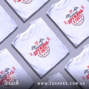 Футболка найкраща дружина. Біла футболка з коротким рукавом, зображена в ракурсі спереду, на фоні світло-сірого кольору. Принт червоного кольору з чорними елементами, напис "Найкраща дружина у світі" оточений декоративними сердечками та візерунками.