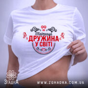 Футболка найкраща дружина. Біла футболка, складена на поверхні, зображена зверху. Принт червоного кольору з чорними елементами, напис "Найкраща дружина у світі" оточений декоративними сердечками та візерунками. На футболці видно етикетку.
