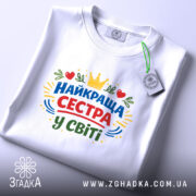 Футболка найкраща сестра, біла, на плоскій поверхні, фон світло-сірий, принт з написом 'НАЙКРАЩА СЕСТРА У СВІТІ' у кольорах червоному, зеленому, жовтому та синьому, з елементами сердець та корони, з біркою на комірці.