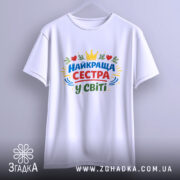 Футболка найкраща сестра, біла, підвішена на вішалці, фон світло-сірий, принт з написом 'НАЙКРАЩА СЕСТРА У СВІТІ' у кольорах червоному, зеленому, жовтому та синьому, з елементами сердець та корони.