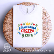 Футболка найкраща сестра, біла, на плоскій поверхні, фон світло-сірий, принт з написом 'НАЙКРАЩА СЕСТРА У СВІТІ' у кольорах червоному, зеленому, жовтому та синьому, з елементами сердець та корони, розташована на плетеній основі.