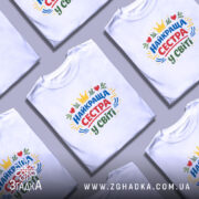 Футболка найкраща сестра, біла, на моделі з коротким рукавом, фон світло-сірий, принт з написом 'НАЙКРАЩА СЕСТРА У СВІТІ' у кольорах червоному, зеленому, жовтому та синьому, з елементами сердець та корони.