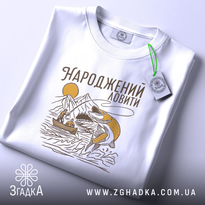 Футболка народжений ловити, біла, з верхнього ракурсу, фон світло-фіолетовий, принт з рибою, горами та човном, виконаний у жовто-коричневих тонах, з етикеткою на зеленому шнурку.