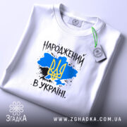 Футболка народжений в Україні, біла, на столі, з фоном у світло-сірому кольорі, складена, принт з жовто-блакитним тризубом на синьому фоні, текст "НАРОДЖЕНИЙ В УКРАЇНІ" виконаний у чорному кольорі.
