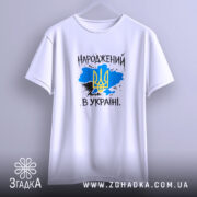 Футболка народжений в Україні, біла, підвішена на вішалці, з фоном у світло-фіолетових тонах, принт з жовто-блакитним тризубом на синьому фоні, текст "НАРОДЖЕНИЙ В УКРАЇНІ" виконаний у чорному кольорі.