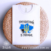 Футболка народжений в Україні, біла, на плетеній кошику, з фоном у світло-сірому кольорі, складена, принт з жовто-блакитним тризубом на синьому фоні, текст "НАРОДЖЕНИЙ В УКРАЇНІ" виконаний у чорному кольорі.