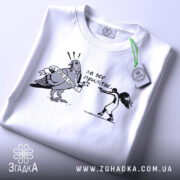 Футболка не будь як голуб. Біла футболка, складена на столі, з принтом чорного кольору, що зображує птаха з банером та дівчину, яка тримає пістолет. Фон світло-сірий, на футболці є етикетка з зеленим шнурком.