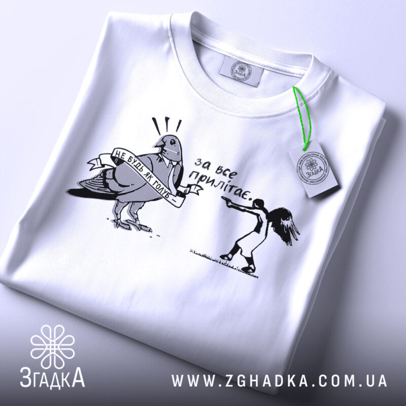 Футболка не будь як голуб. Біла футболка, складена на столі, з принтом чорного кольору, що зображує птаха з банером та дівчину, яка тримає пістолет. Фон світло-сірий, на футболці є етикетка з зеленим шнурком.