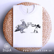 Футболка не будь як голуб. Біла футболка, складена на плетеному кошику, з принтом чорного кольору, що зображує птаха з банером та дівчину, яка тримає пістолет. Фон світло-сірий, видима етикетка на комірці.