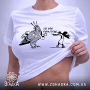 Футболка не будь як голуб. Біла футболка, складена на столі, з принтом чорного кольору, що зображує птаха з банером та дівчину, яка тримає пістолет. Фон світло-сірий, на футболці є етикетка з зеленим шнурком.