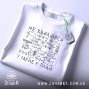 Футболка не хвилюйся у мене є план. Біла футболка, складена на поверхні, з видимим принтом. На фоні світло-сірий колір. Принт чорного кольору з математичними формулами та написом 'НЕ ХВИЛЮЙСЯ У МЕНЕ Є ПЛАН'.