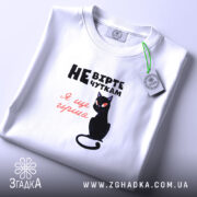 Футболка не вірте чуткам, біла, складена на поверхні, фон світло-фіолетовий, принт з чорним котом та текстом у чорному і червоному кольорах.