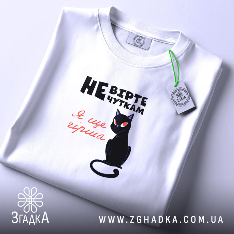 Футболка не вірте чуткам, біла, складена на поверхні, фон світло-фіолетовий, принт з чорним котом та текстом у чорному і червоному кольорах.