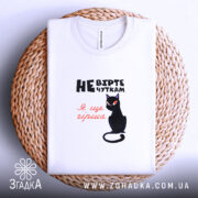 Футболка не вірте чуткам, біла, складена на плетеній основі, фон світло-фіолетовий, принт з чорним котом та текстом у чорному і червоному кольорах.