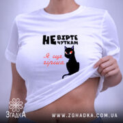 Футболка не вірте чуткам, біла, складена на поверхні, фон світло-фіолетовий, принт з чорним котом та текстом у чорному і червоному кольорах.