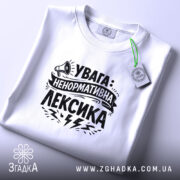 Футболка ненормативна лексика, біла, на столі, фон світло-сірий, принт чорний з написом 'УВАГА: НЕНОРМАТИВНА ЛЕКСИКА' та малюнком гучномовця, з етикеткою.