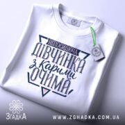 Футболка непосидюча дівчинка, білого кольору, зображена в складеному вигляді на світлому фоні, принт на футболці містить текст "непосидюча дівчинка з карими очима" в темно-синіх та рожевих відтінках, з етикеткою, що підвішена зеленим шнурком.