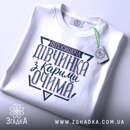 Футболка непосидюча дівчинка, білого кольору, зображена в складеному вигляді на світлому фоні, принт на футболці містить текст "непосидюча дівчинка з карими очима" в темно-синіх та рожевих відтінках, з етикеткою, що підвішена зеленим шнурком.