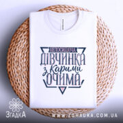 Футболка непосидюча дівчинка, білого кольору, зображена в складеному вигляді на плетеній кошику, принт на футболці містить текст "непосидюча дівчинка з карими очима" в темно-синіх та рожевих відтінках.