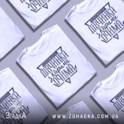 Футболка непосидюча дівчинка, білого кольору, зображена з близького ракурсу, на фоні світло-фіолетового кольору, принт на футболці містить текст "непосидюча дівчинка з карими очима" в темно-синіх та рожевих відтінках.