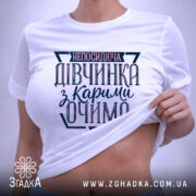 Футболка непосидюча дівчинка, білого кольору, зображена в складеному вигляді на світлому фоні, принт на футболці містить текст "непосидюча дівчинка з карими очима" в темно-синіх та рожевих відтінках, з етикеткою, що підвішена зеленим шнурком.