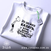 Футболка ну шо його казать, біла футболка, складена на поверхні, з принтом, що містить персонажа з сумним виразом обличчя та текстом "Ну шо його казать, коли воно канешно". На футболці є етикетка з зеленим шнурком. Фон світло-сірий.