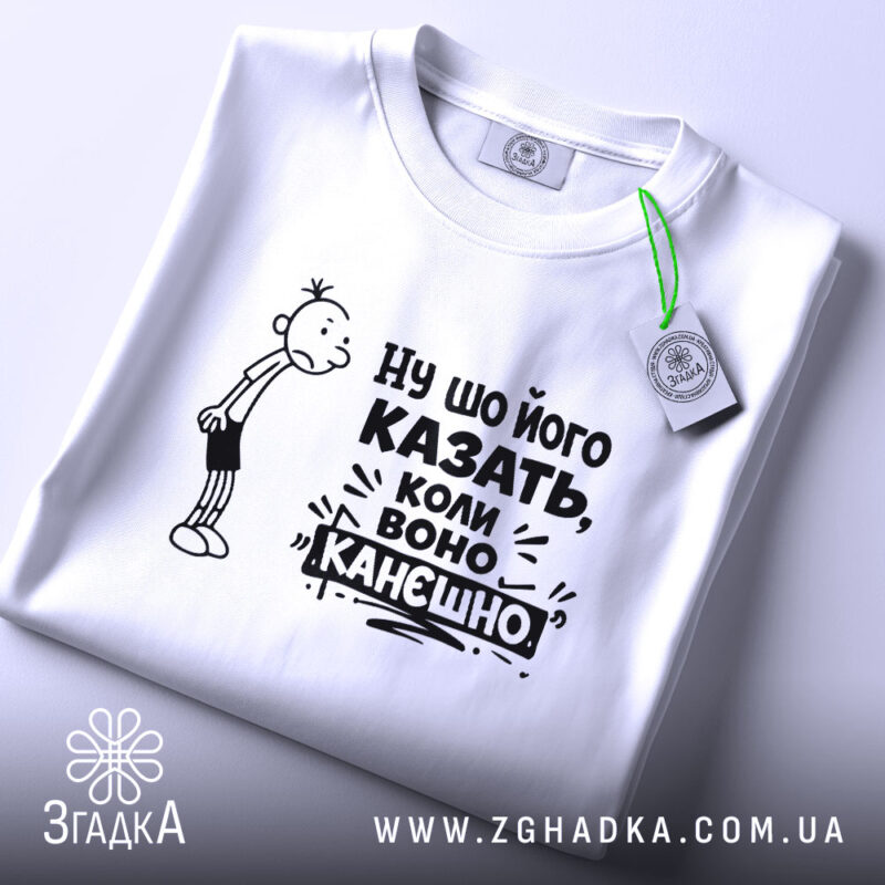 Футболка ну шо його казать, біла футболка, складена на поверхні, з принтом, що містить персонажа з сумним виразом обличчя та текстом "Ну шо його казать, коли воно канешно". На футболці є етикетка з зеленим шнурком. Фон світло-сірий.
