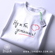 Футболка ну ж бо усміхнись, складена біла футболка, ракурс зверху, фон світло-сірий, принт з написом 'Ну ж бо, усміхнись!' у чорному кольорі з червоним сердечком, на ярлику зелена стрічка.