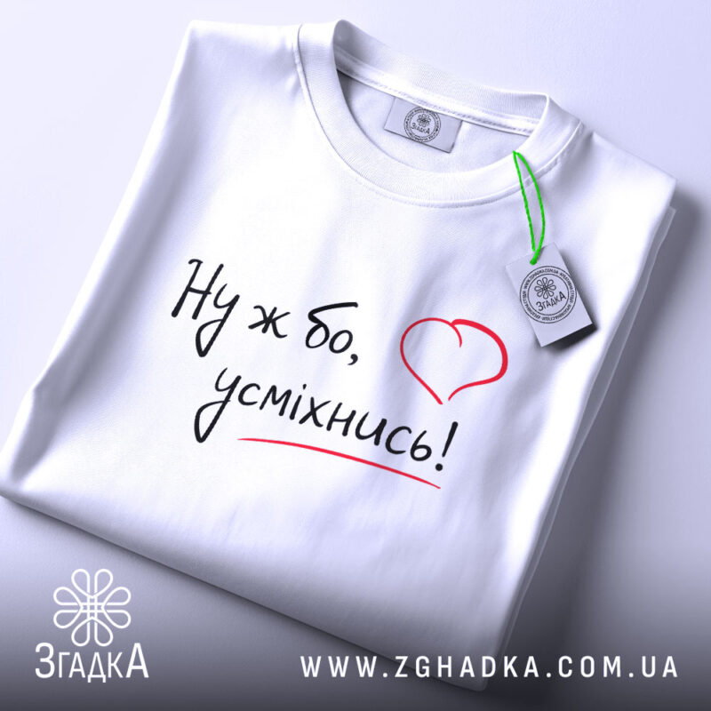 Футболка ну ж бо усміхнись, складена біла футболка, ракурс зверху, фон світло-сірий, принт з написом 'Ну ж бо, усміхнись!' у чорному кольорі з червоним сердечком, на ярлику зелена стрічка.