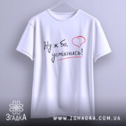 Футболка ну ж бо усміхнись, біла футболка на вішалці, ракурс спереду, фон світло-сірий, принт з написом 'Ну ж бо, усміхнись!' у чорному кольорі з червоним сердечком.