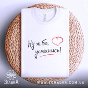 Футболка ну ж бо усміхнись, складена біла футболка на плетеній тарілці, ракурс зверху, фон світло-сірий, принт з написом 'Ну ж бо, усміхнись!' у чорному кольорі з червоним сердечком.