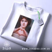 Футболка-оберіг від ідіотів, біла футболка, розкладена на поверхні, з принтом жінки, що формує серце руками, на фоні світлого кольору, з етикеткою, що звисає з коміра.