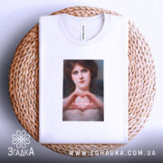 Футболка-оберіг від ідіотів, біла футболка, складена на плетеній кошику, з принтом жінки, що формує серце руками, на фоні світлого кольору.