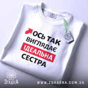 Футболка ось так виглядає ідеальна сестра. Біла футболка, складена на столі, з видимим принтом. На фоні світлий однотонний колір. Принт містить червоний текст на чорному фоні, що говорить: 'Ось так виглядає ідеальна сестра', а також зелена етикетка з логотипом.